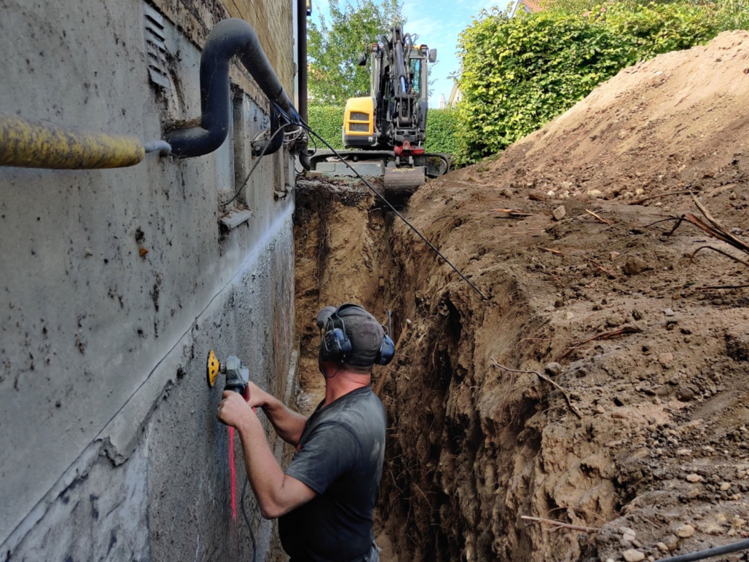 Hantverkare slipar grundmur i dräneringsschakt med grävmaskin i bakgrunden